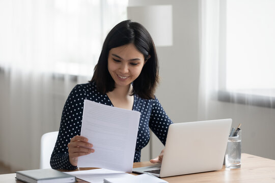 Happy millennial Asian girl sit at desk work on laptop consider read good news in post paper correspondence or letter document, smiling Vietnamese young woman study on computer prepare report