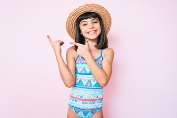 Young little girl with bang wearing swimsuit and summer hat smiling and looking at the camera pointing with two hands and fingers to the side.