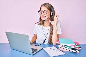 Young blonde girl wearing operator headset at the call center office smiling with hand over ear listening and hearing to rumor or gossip. deafness concept.