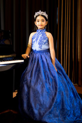 Fototapeta premium A portrait of a cute Asian girl, makeup, wearing a blue evening dress and a crown standing beside the piano in the room.