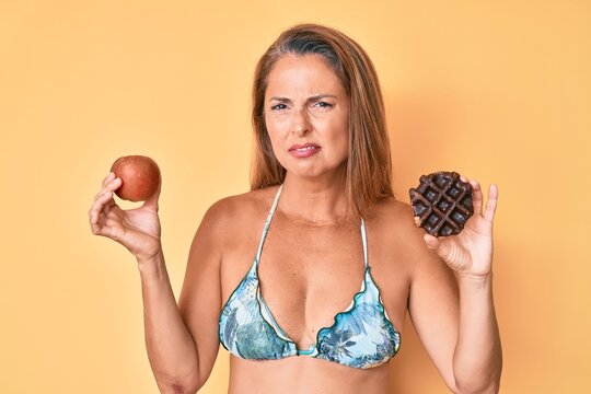Middle age hispanic woman holding apple and donut clueless and confused expression. doubt concept.
