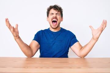 Young caucasian man with curly hair wearing casual clothes sitting on the table crazy and mad...