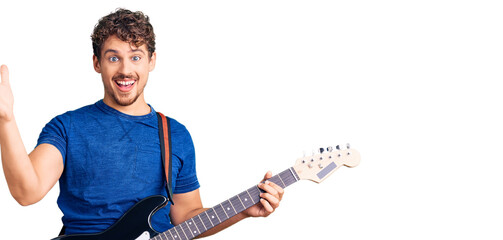 Young handsome man with curly hair playing electric guitar celebrating victory with happy smile and winner expression with raised hands