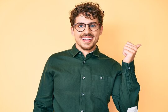 Young caucasian man with curly hair wearing casual clothes and glasses smiling with happy face looking and pointing to the side with thumb up.