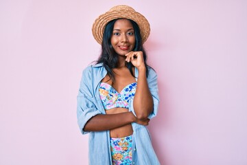 Young indian girl wearing bikini and summer hat smiling looking confident at the camera with crossed arms and hand on chin. thinking positive.