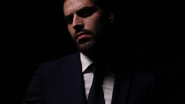 young elegant businessman in suit posing in a fashion light, looking to side and adjusting tie and suit, crossing arms and holding hand to face touching lips and chin, thinking and standing