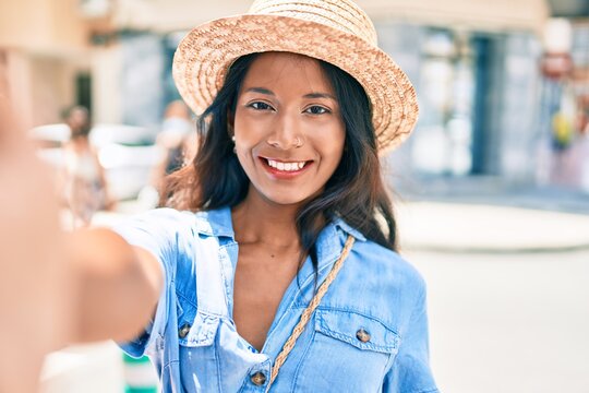 Young beautiful indian woman wearing summer hat smiling happy making selfie by the camera at the city.