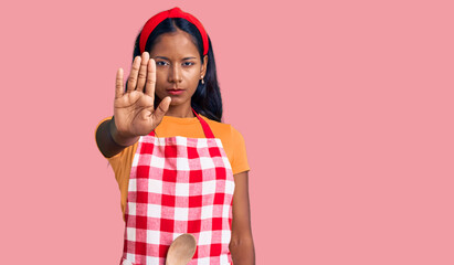 Young indian girl wearing professional baker apron doing stop sing with palm of the hand. warning expression with negative and serious gesture on the face.