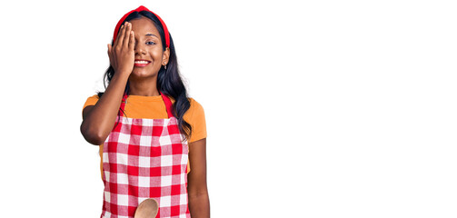 Young indian girl wearing professional baker apron covering one eye with hand, confident smile on...