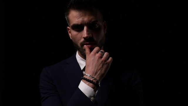 sensual young businessman in suit touching lips and admiring, moving head and analyzing on dark background