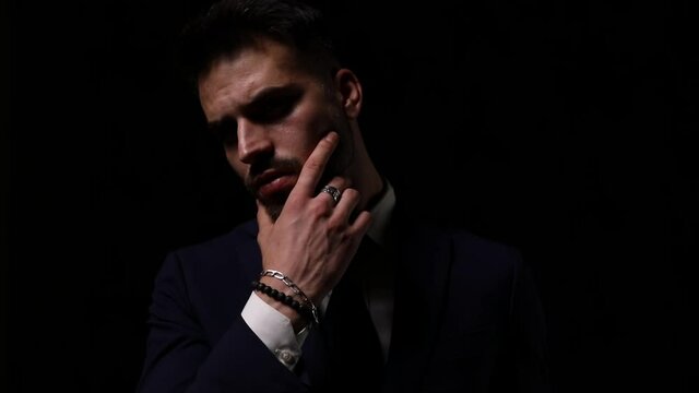 sexy young man in suit holding hand to face and touching chin and beard, arranging suit and moving on dark background