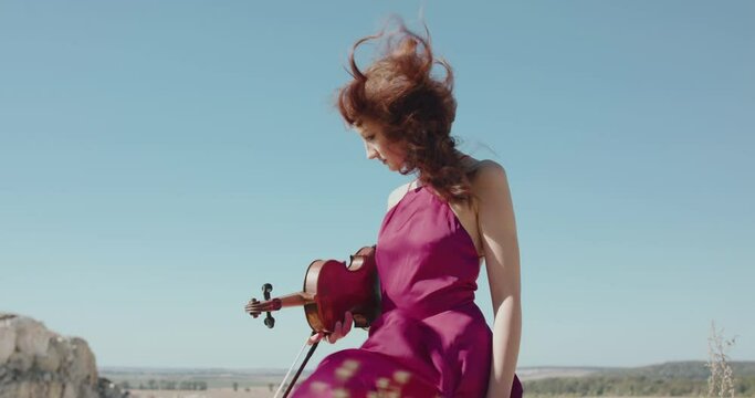 Girl with blowing long hair and windy dress stands with violin in hand on cliff