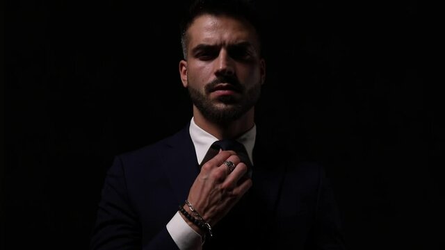sexy young businessman adjusting tie and suit, frowning, moving in a side view pose and walking away on dark background