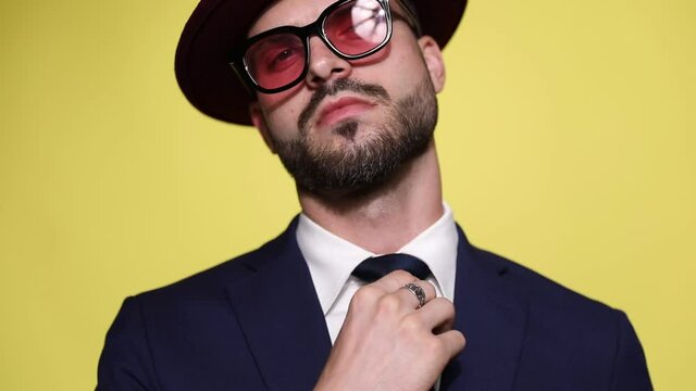 elegant young businessman wearing sunglasses and hat, fixing tie and suit, crossing arms and smirking,smiling and leaving on yellow background