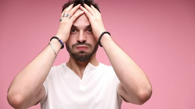 sexy casual man wearing a white T-shirt, looking at camera, running his fingers trough his head, puffing cheeks, covering his mouth, thinking and being anxious on pink background