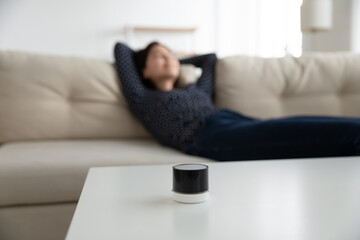 Focus on mini modern smart wireless speaker on table, woman relax rest on sofa in living room at background, female listen to music good quality sound loudspeaker audio device on couch