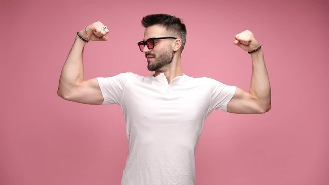 attractive macho casual man is flexing his muscles, kissing his biceps with pride, bowing his head and wearing sunglasses on pink background