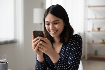 Happy millennial Asian girl look at smartphone screen browse surf wireless internet in gadget,...