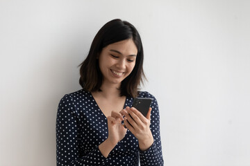 Happy millennial Vietnamese girl stand isolated on grey studio background use modern cellphone...