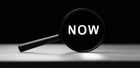 Magnifying glass with text NOW on wooden table. Concept of search