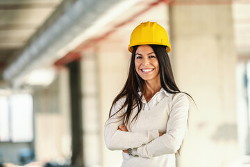 Young smiling female architect standing in building in construction process with arms crossed and looking at camera.