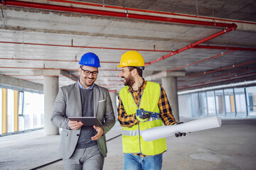 Smiling architect in suit with helmet on head talking to contractor while standing in building in...