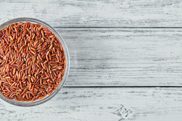 Fototapeta na wymiar Raw red rice grains in a transparent glass plate on wooden background. Closeup, top view, copy space