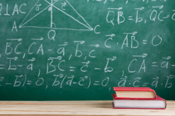 Education and reading concept - group of books on the wooden table, green blackboard with formulas on the background. Teachers desk