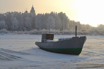 Winter landscape with a fishing boat on the shore of the frozen bay against the frosty sky, groves and domes of the church, Rybinsk, Russia