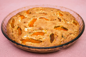 A large, round baked apple and apricot pie stands in a glass form on a table with a tablecloth.