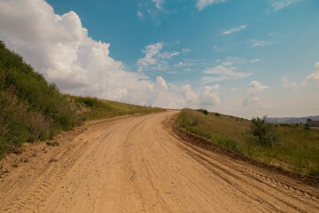 Scenic beautiful nature with green grass and dramatic cloudy sky in hot summer day. Yellow sandy road and spirit of adventures, mountains on a background