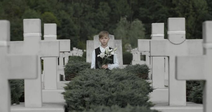 Portrait of teenger boy looking to camera while standing in row of stone crosses. Young kid holding white lily flower at cemetery. Concept of memorial day