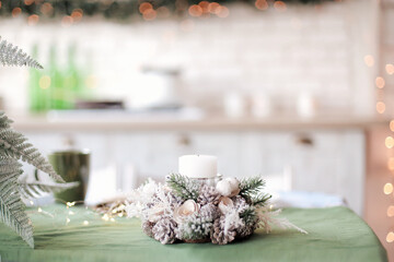 Christmas decorations. Home interior in pastel colors. Holiday candle decorated with pine cones and snow fir branches. Multi-colored bokeh in the background. Selective focuse. Copy space for text