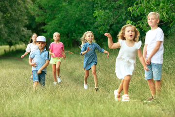 Kids, children running on meadow in summer's sunlight. Look happy, cheerful with sincere bright emotions. Cute caucasian boys and girls. Concept of childhood, happiness, movement, family and summer.