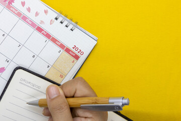 A woman's hand holding a pen writing on the notebook placed on the calendar. There are a copy space. Business or travel trip concept.