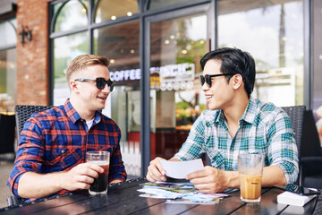 Smiling handsome young men in sunglasses sitting at cafe table and discussing printed photos from summer trip