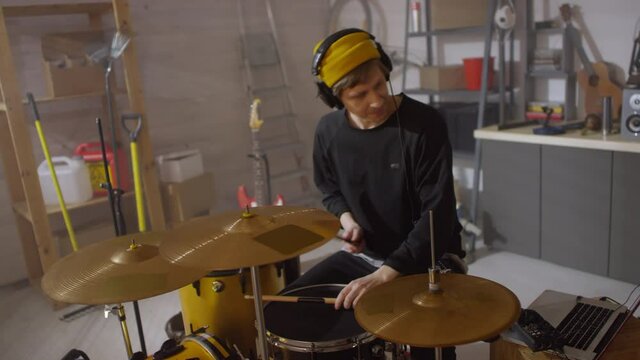 Young Caucasian man wearing casual outfit putting on headphones and starting playing drums in garage studio