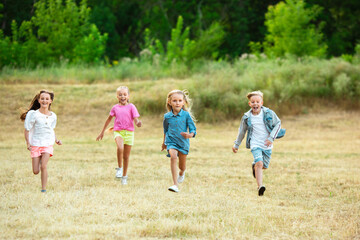 Fototapeta premium Kids, children running on meadow in summer's sunlight. Look happy, cheerful with sincere bright emotions. Cute caucasian boys and girls. Concept of childhood, happiness, movement, family and summer.