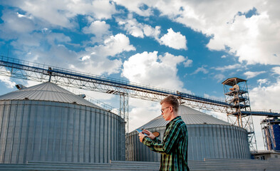 Naklejka na ściany i meble A young guy stands with a tablet near a metal elevator in an agricultural area. A grain warehouse.