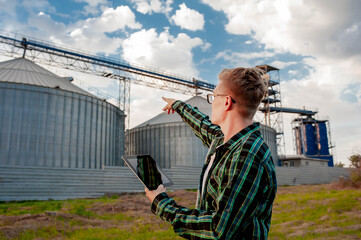  A young guy stands with a tablet near a metal elevator in an agricultural area. A grain warehouse. © александр таланцев