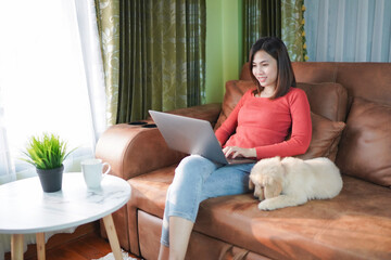 Happy asian woman with puppy at home and relaxing with laptop computer on the sofa in the living room. stay at home for coronavirus prevention campaign.Working from home and Online shopping concept.