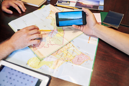 Man pointing at coast map when his friend showing photos from this place on smartphone