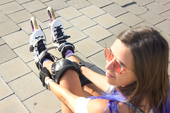 Young woman with blue hair and sunglasses putting on roller skates and tie the shoelaces outdoors. Summertime sport activity concept.