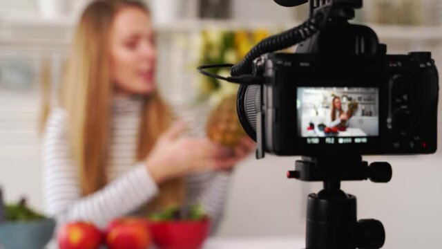 Camera screen recording - focus transition - Food blogger cooking fresh vegan salad of fruits in kitchen studio, filming tutorial. Female influencer holds apple, pineapple, talks about healthy eating.