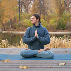 Young pretty woman doing yoga poses in the park autumn next to the lake shore with yellow grass ....