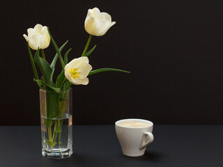 Bouquet of yellow tulips in vase with cup on a black background.