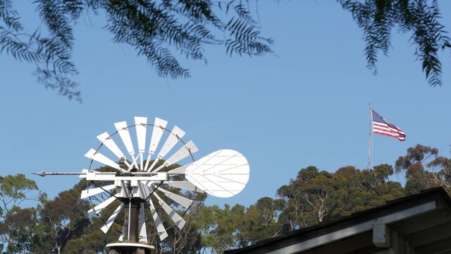 Classic retro windmill, bladed rotor and USA flag against blue sky. Vintage water pump wind turbine, power generator on livestock ranch or agricultural farm. Rural symbol of wild west, rustic suburb