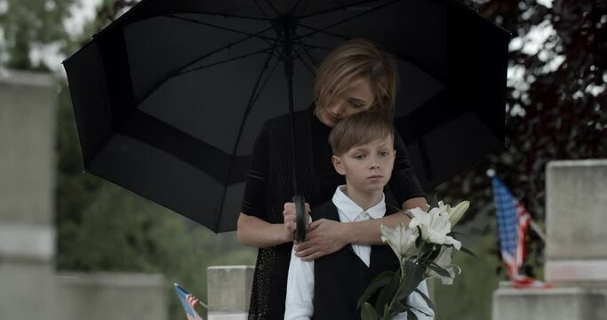 Sad woman hugging and leaned to his teen kid while standing under umbrella at cemetery.Widow and young boy with white flowers near gravestone with american flag.Concept of memorial day