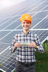 Portrait of happy man with hundred dollar bill in hand shows thumb a sign like he likes in the background of a solar station. Business and generation concept. Green ecological power energy generation.