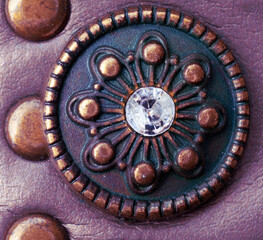 Macro shot of a copper medaillon with flower pattern and a light reflection on the glass bead in the centre. Fake purple leather background with three more buttons on the left side of the frame.    